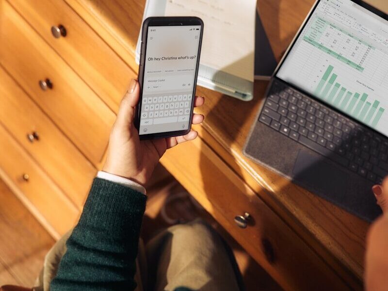 Person holding phone near laptop and tablet on desk
